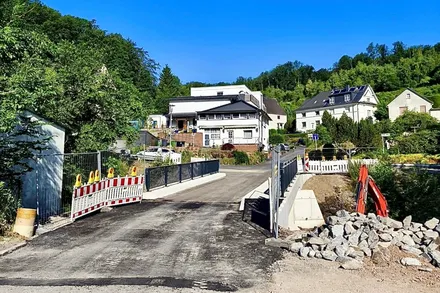 Die neue Brücke über die Volme ist für den Verkehr freigegeben, nachdem die alte Brücke an der „Schiefen Ahelle“ ersetzt wurde. Im Vordergrund sind Absperrungen und Bauarbeiten zu sehen, im Hintergrund Wohnhäuser und Bäume.