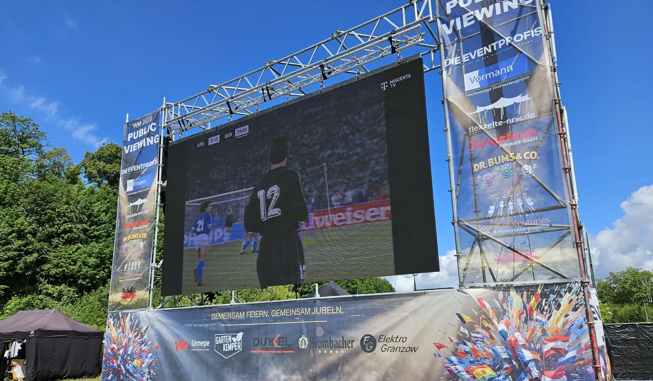 Große LED-Wand zeigt Fußballspieler mit der Nummer 12 während der Vorbereitungen für eine WM-Party. Werbebanner für Sponsoren umrahmen die Leinwand.