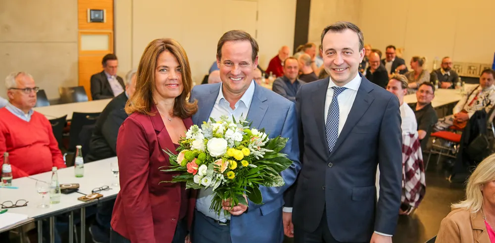 Drei Personen lächeln in die Kamera: Nicola Nähle, Thorsten Schick, der einen Blumenstrauß hält, und Paul Ziemiak. Im Hintergrund sind weitere Personen und Tische in einem Konferenzraum zu sehen.