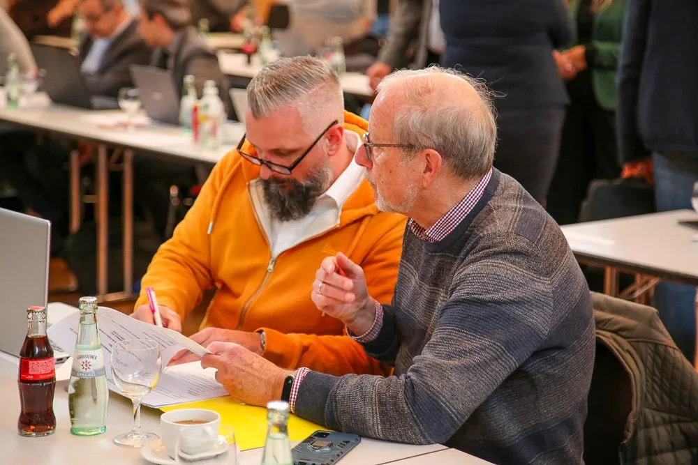 Nils Koschinsky und Detlef Köpke von der FDP-Gruppe diskutieren bei einer Veranstaltung an einem Tisch mit Unterlagen, Getränken und einem Laptop.
