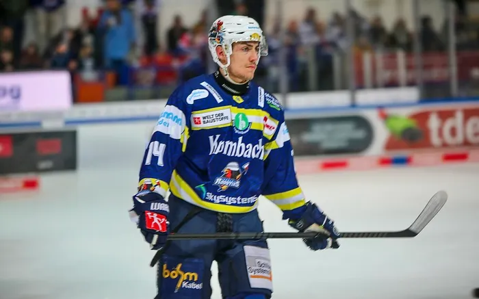 Eishockeyspieler Noel Saffran in voller Ausrüstung mit Schläger auf dem Eis, während eines Spiels. Sein Trikot zeigt verschiedene Werbesponsoren.