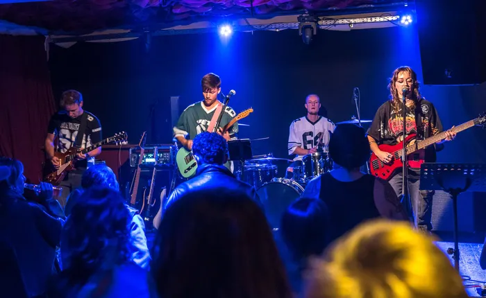 Eine Rockband spielt mit Gitarren und Schlagzeug unter blauem Bühnenlicht vor einem Publikum bei einem Benefizkonzert.