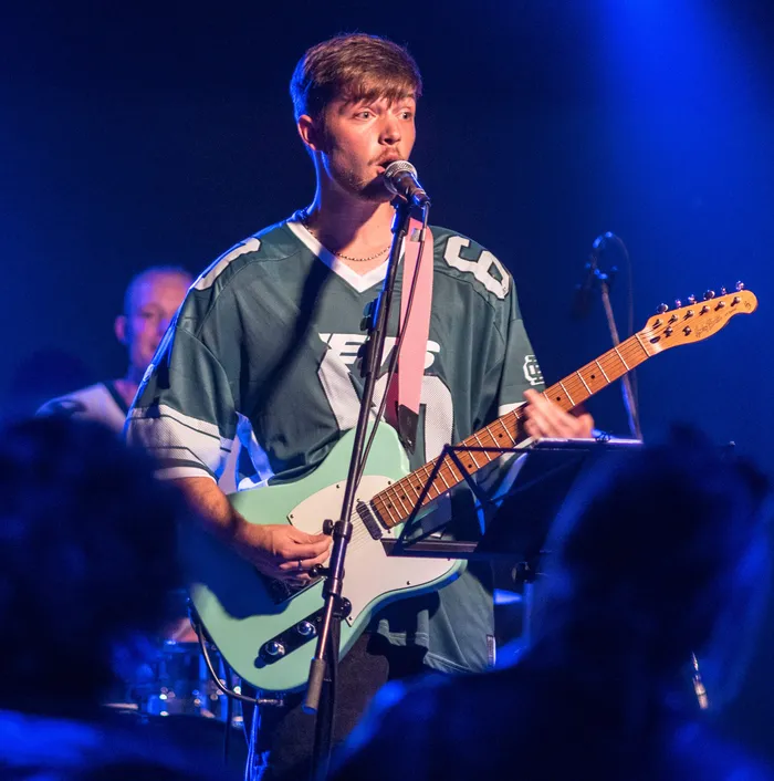 Ein Musiker in einem Sporttrikot singt in ein Mikrofon und spielt eine hellblaue E-Gitarre bei einem Benefizkonzert.