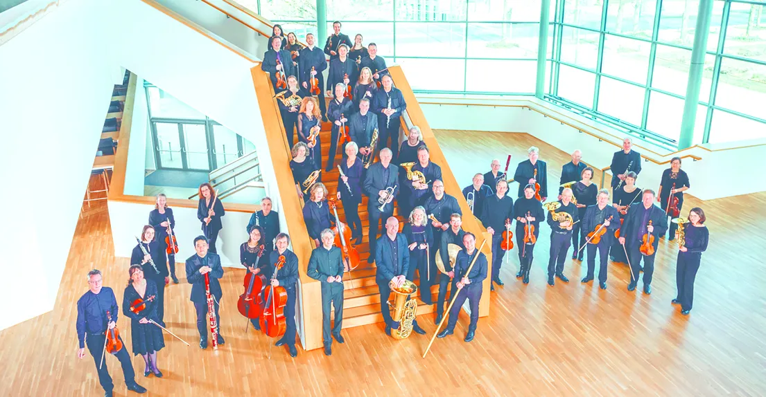 Das Städtische Orchester Osnabrück posiert auf einer breiten Treppe in einem modernen Gebäude mit Glasfassade. Die Musikerinnen und Musiker spielen verschiedene Instrumente.