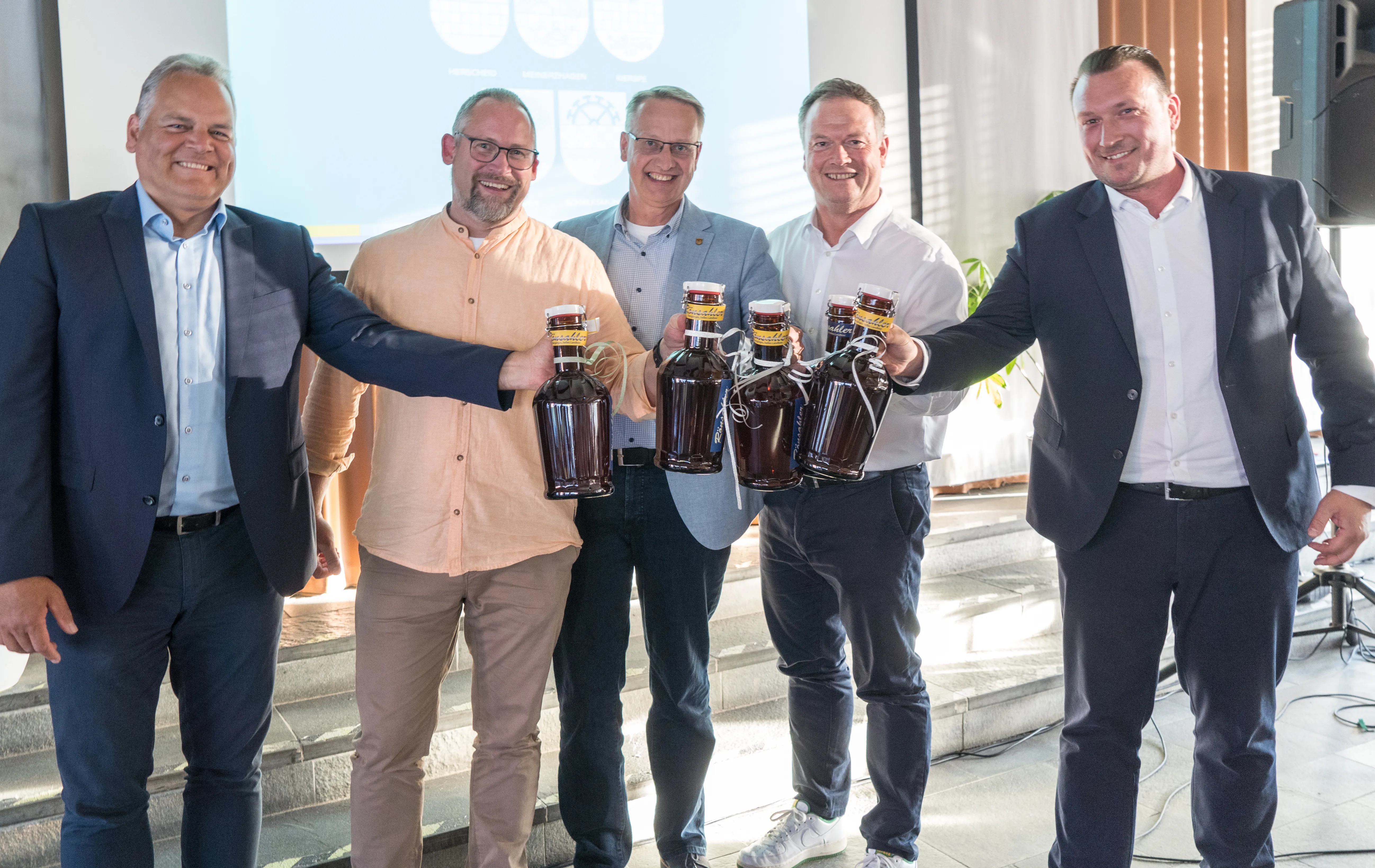 Fünf Männer halten jeweils eine Flasche dunkles Getränk in die Höhe, lächeln in die Kamera. Im Hintergrund ist ein Wappen zu sehen.