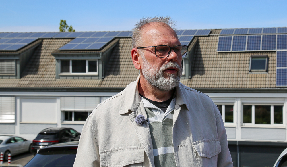 Mann mit Brille und Bart, vor dem Hintergrund eines Gebäudes mit Solaranlagen. Er trägt ein Mikrofon und blickt in die Kamera.