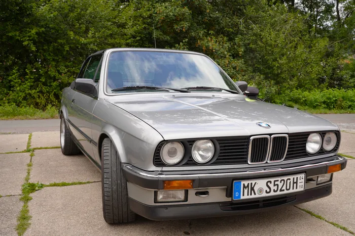 Ein silberner BMW 325i steht auf einem gepflasterten Platz vor grünen Bäumen.