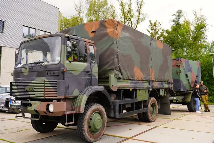Ein olivgrün-braun getarnter Iveco-LKW mit abgedeckter Ladefläche und einem passenden Anhänger auf einem gepflasterten Platz.