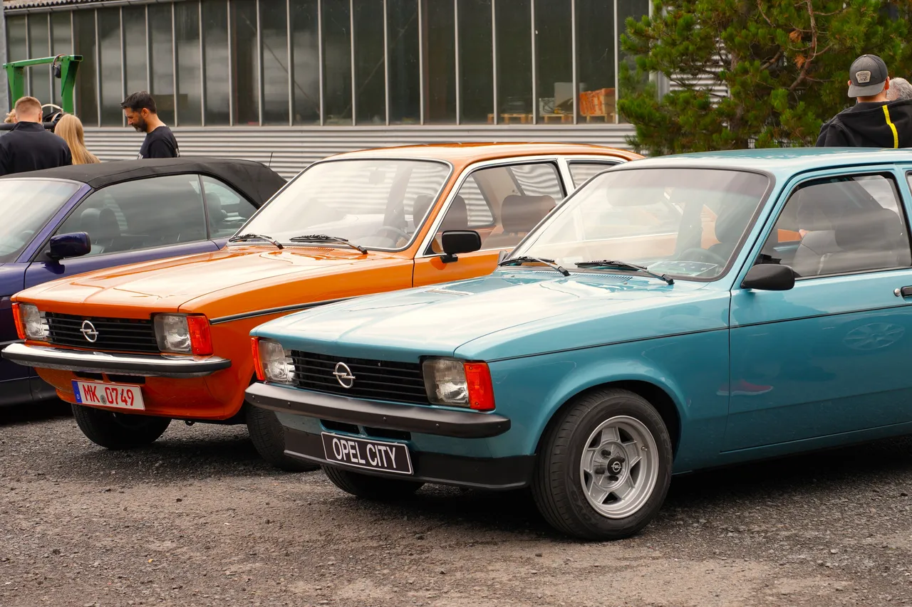 Ein hellblauer Opel City, ein orangefarbenes Auto und ein dunkles Fahrzeug stehen nebeneinander auf einem Schotterplatz bei einem Oldtimer-Treffen.