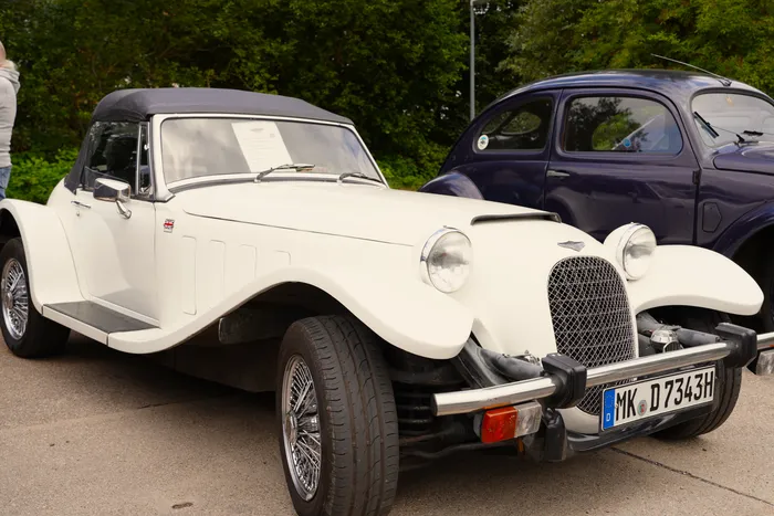 Ein weißes Oldtimer-Cabriolet mit schwarzem Verdeck steht neben einem dunkelblauen Oldtimer.