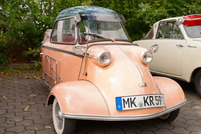 Ein pfirsichfarbenes Oldtimer-Kleinstauto mit Glaskuppel steht auf einem gepflasterten Platz neben einem weißen Fahrzeug.