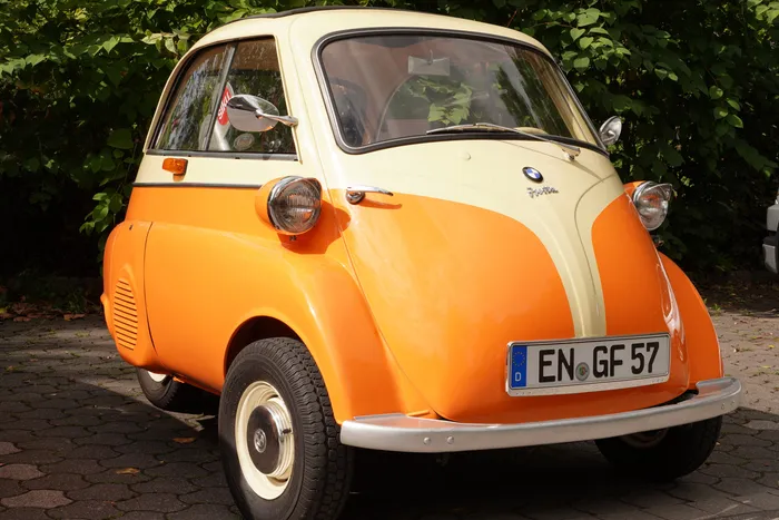 Ein kleiner, zweifarbiger Oldtimer in Orange und Creme steht auf einem gepflasterten Platz vor grünen Sträuchern.