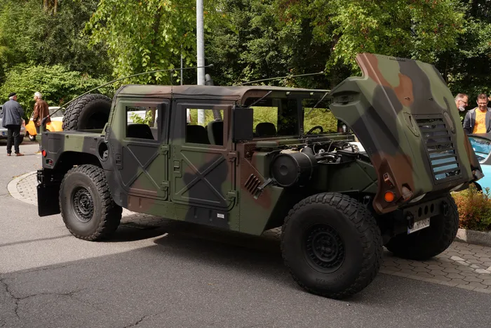 Ein olivgrün-braun getarntes Militärfahrzeug mit geöffneter Motorhaube steht auf einem asphaltierten Platz.