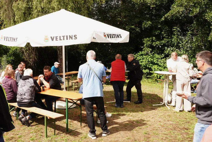 Menschen sitzen an Picknicktischen unter einem großen weißen Veltins-Sonnenschirm im Freien.