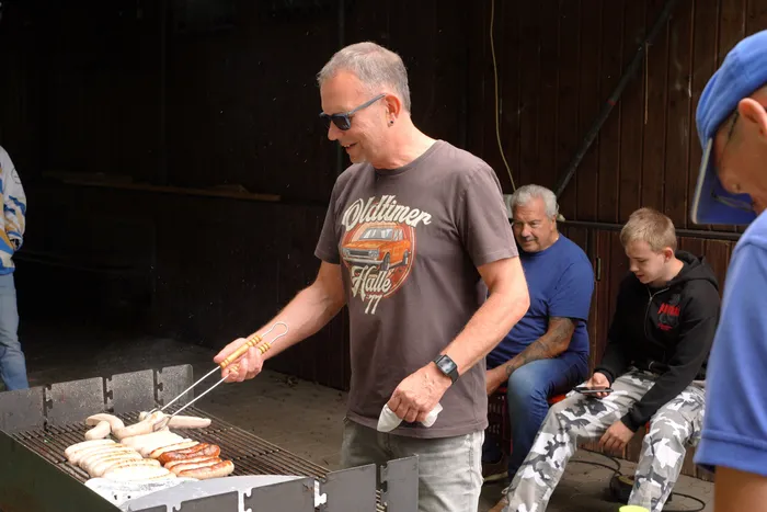Ein Mann wendet mit einer Grillzange Würstchen auf einem Grill bei einem Oldtimer-Treffen.