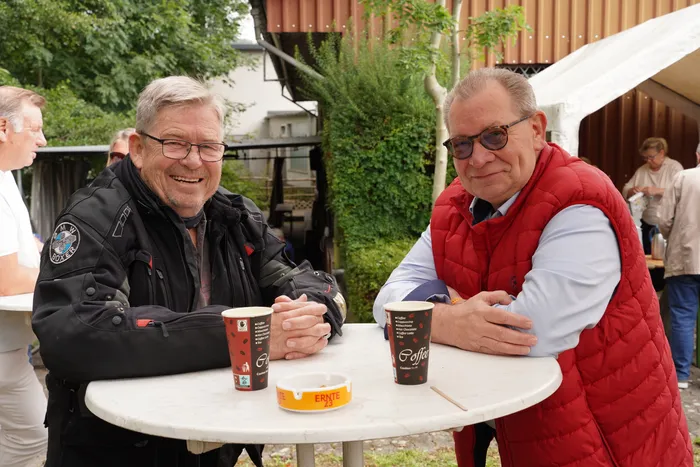 Zwei Männer, einer in Motorradkleidung, sitzen an einem Tisch mit Kaffeebechern bei einem Old- und Youngtimer-Treffen im Freien.