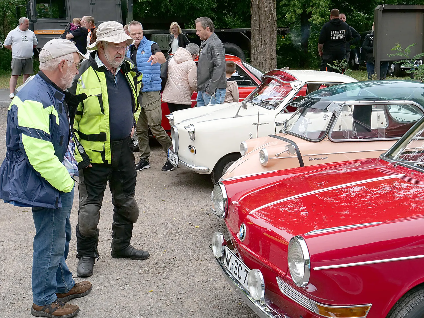 Zwei Männer betrachten einen roten Oldtimer mit geöffneter Motorhaube, während im Hintergrund weitere Oldtimer und Besucher zu sehen sind.
