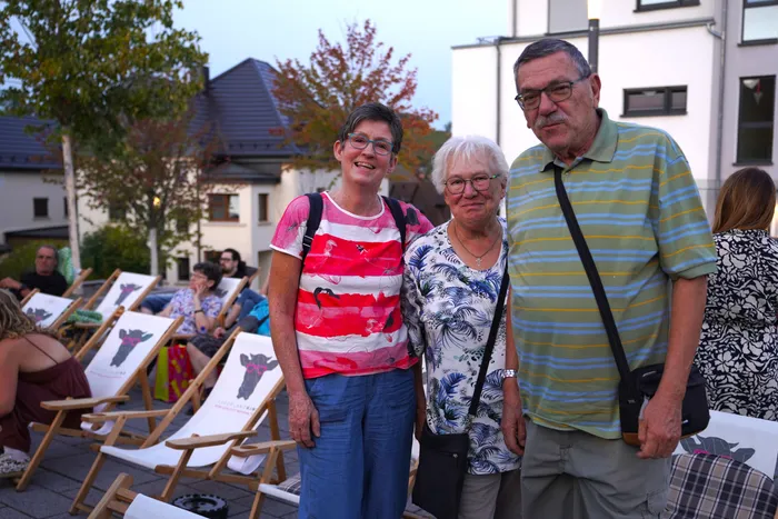 Birgit Baureis und Jeanne Martin stehen lächelnd mit einem Mann vor einer Reihe von Liegestühlen bei einem Open-Air-Kino in Herscheid.
