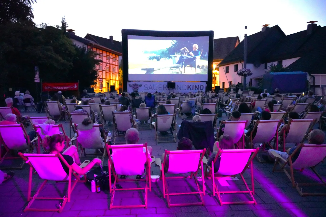 Zuschauer sitzen in Liegestühlen vor einer großen Leinwand bei einem Open-Air-Kino in der Abenddämmerung.