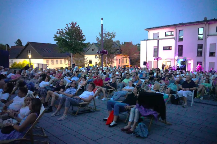 Ein Publikum sitzt in Klappstühlen bei einem Open-Air-Kino auf einem gepflasterten Platz vor beleuchteten Gebäuden in der Abenddämmerung.