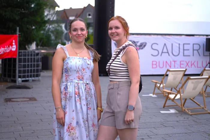 Zwei Frauen, eine im Blumenkleid und eine im gestreiften Top, stehen vor einem Banner bei einer Open-Air-Kinoveranstaltung.