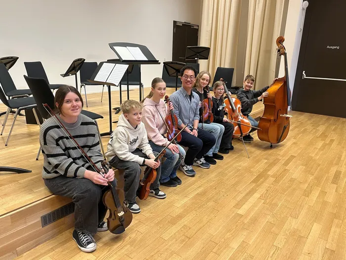 Eine Gruppe von jungen Musikern mit Geigen, Bratschen und einem Cello posiert mit ihrem Dirigenten Shih-Yu Chu in einem hellen Übungsraum.