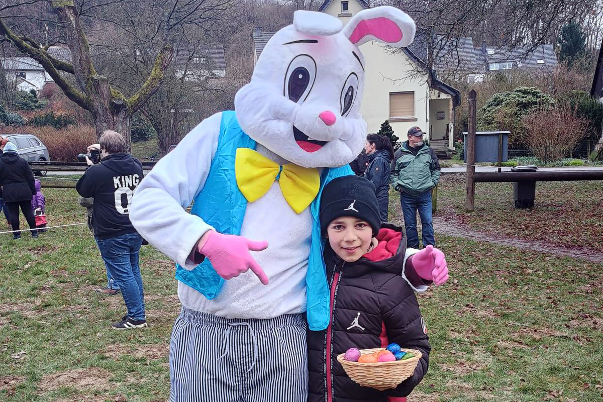 Ein Kind mit einem Korb voller bunter Ostereier steht neben einer Person in einem Hasenkostüm. Im Hintergrund sind weitere Besucher und Gebäude zu sehen.