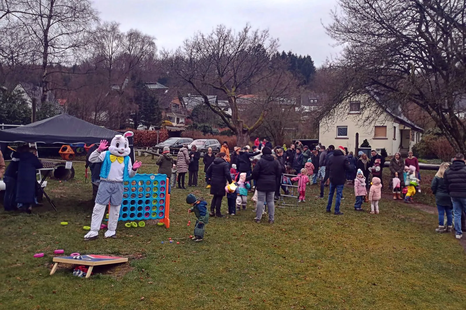 Menschengruppe bei einer Ostereiersuche auf einer Wiese, im Hintergrund Häuser und Bäume. Ein Osterhase in Menschengröße steht neben einem Connect-Four-Spiel.