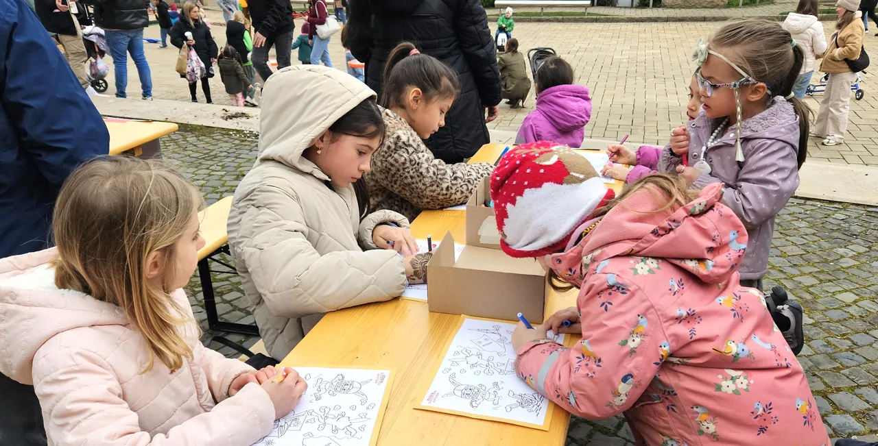 Mehrere Kinder sitzen an einem Tisch im Freien und malen Ostereier auf Papier. Im Hintergrund sind weitere Menschen und ein Spielmobil zu sehen.
