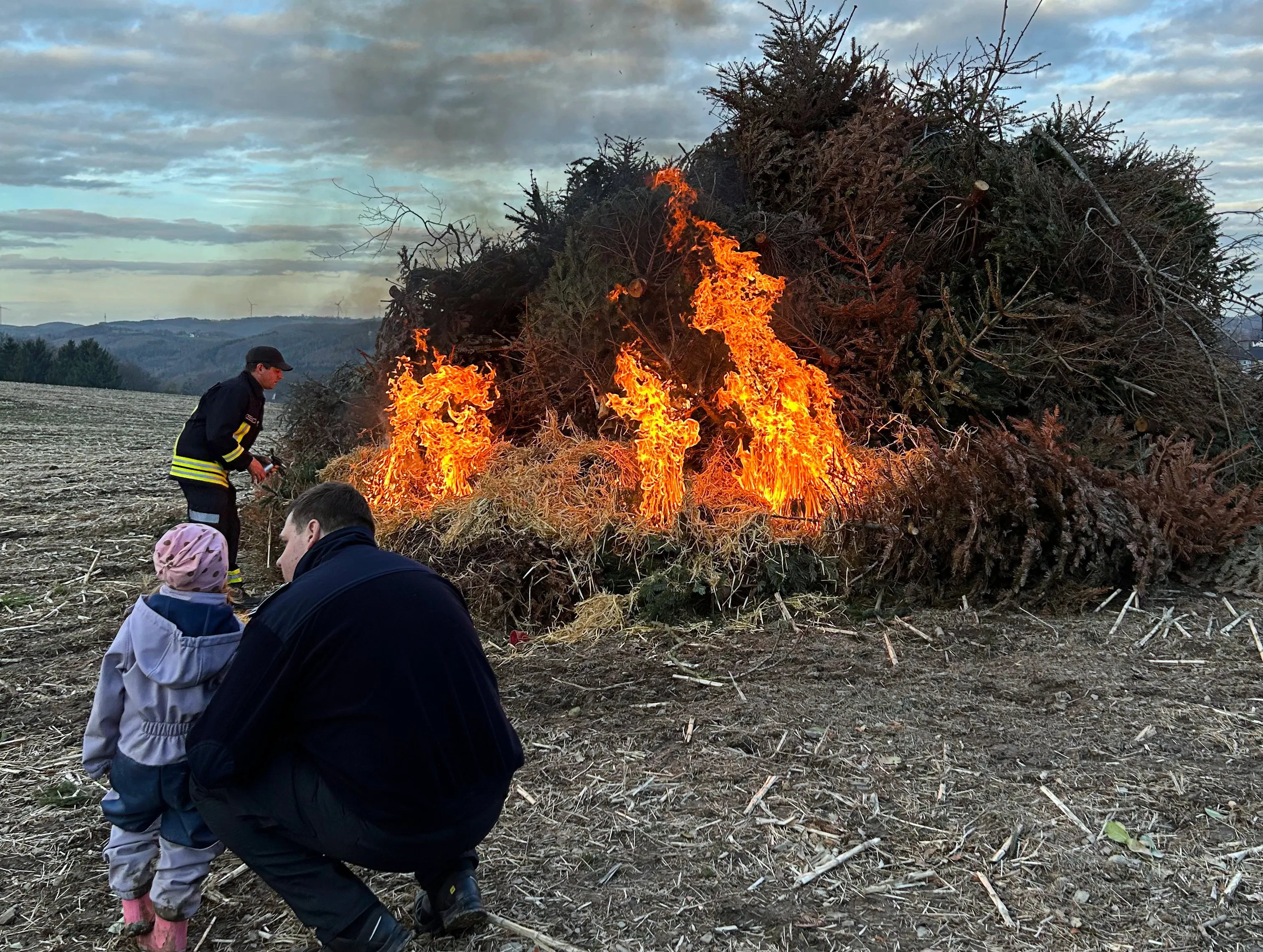 Großes Osterfeuer mit Flammen und Rauch auf einem Feld, umgeben von Zuschauern, darunter ein Kind mit rosa Mütze und ein Mann mit gelber Warnweste.