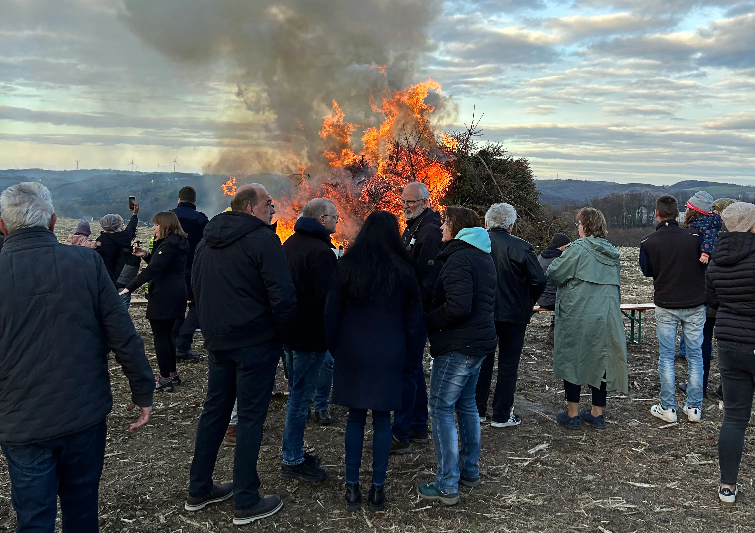 Eine Gruppe Menschen steht um ein großes Osterfeuer mit hellen Flammen und viel Rauch. Im Hintergrund sind Hügel zu sehen.