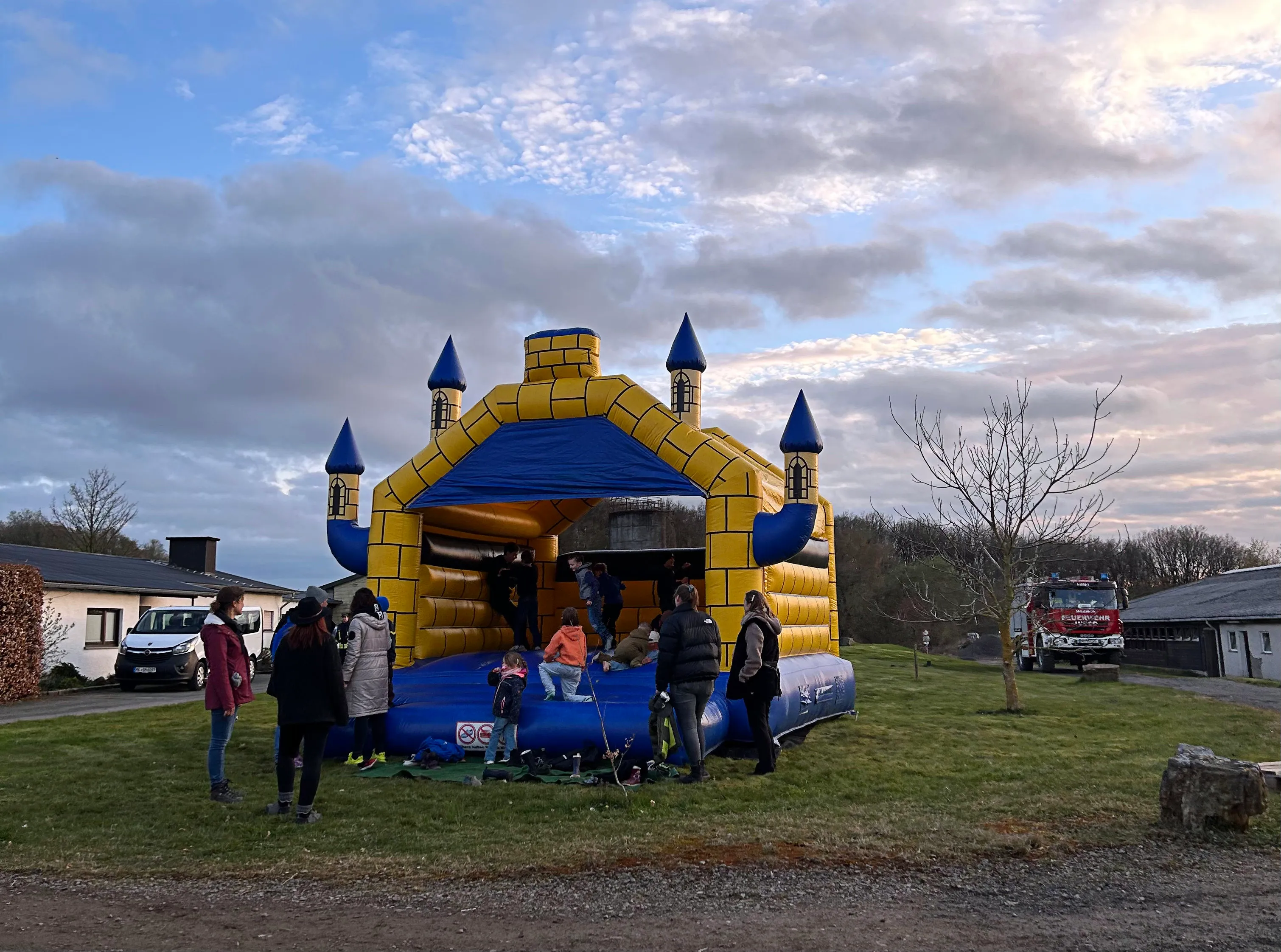 Gelb-blaue Hüpfburg steht auf einer Wiese, umgeben von Menschen. Im Hintergrund sind Gebäude und ein Feuerwehrauto zu sehen.