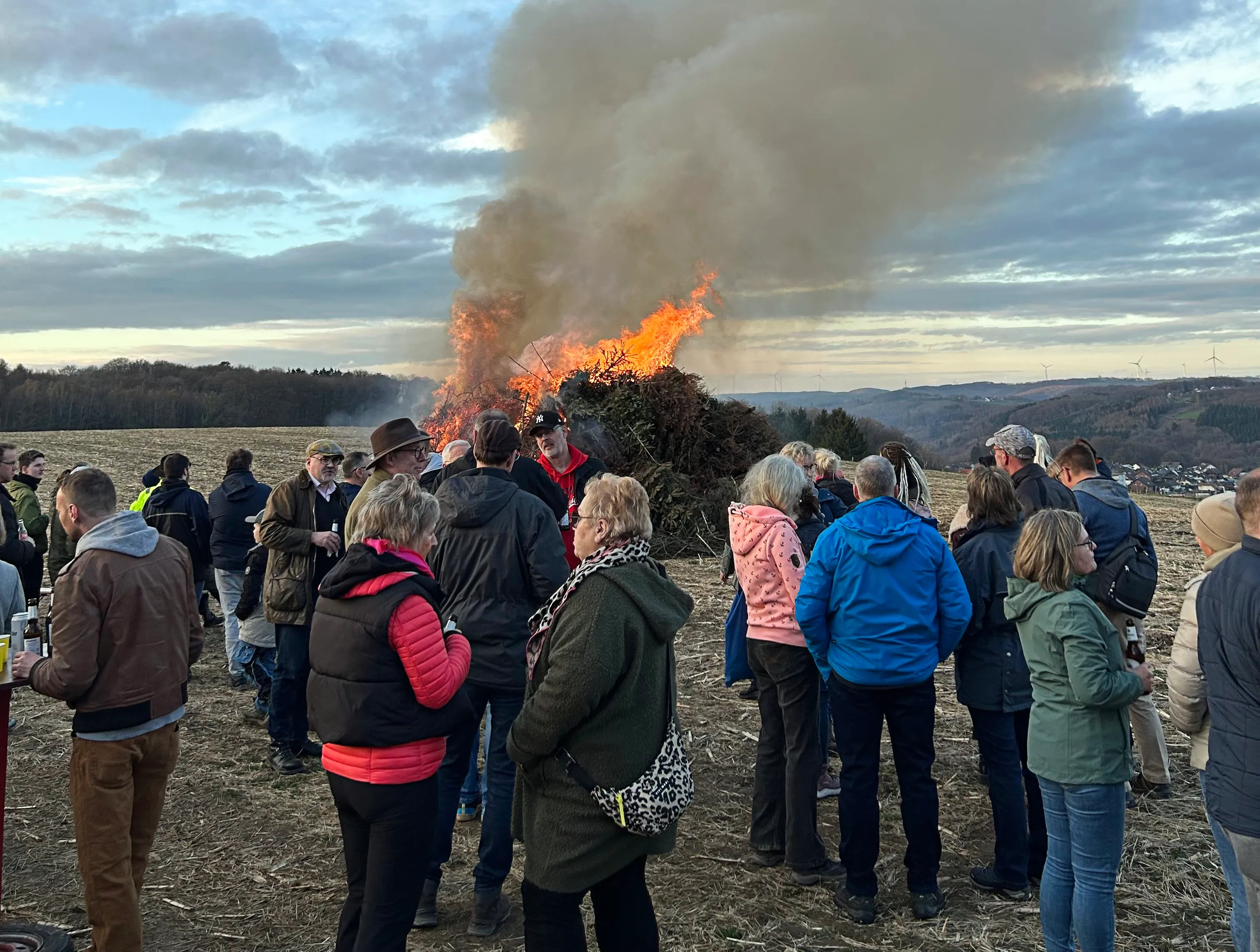 Eine große Menschenmenge beobachtet ein brennendes Osterfeuer auf einem Feld. Rauch steigt in den Himmel auf.