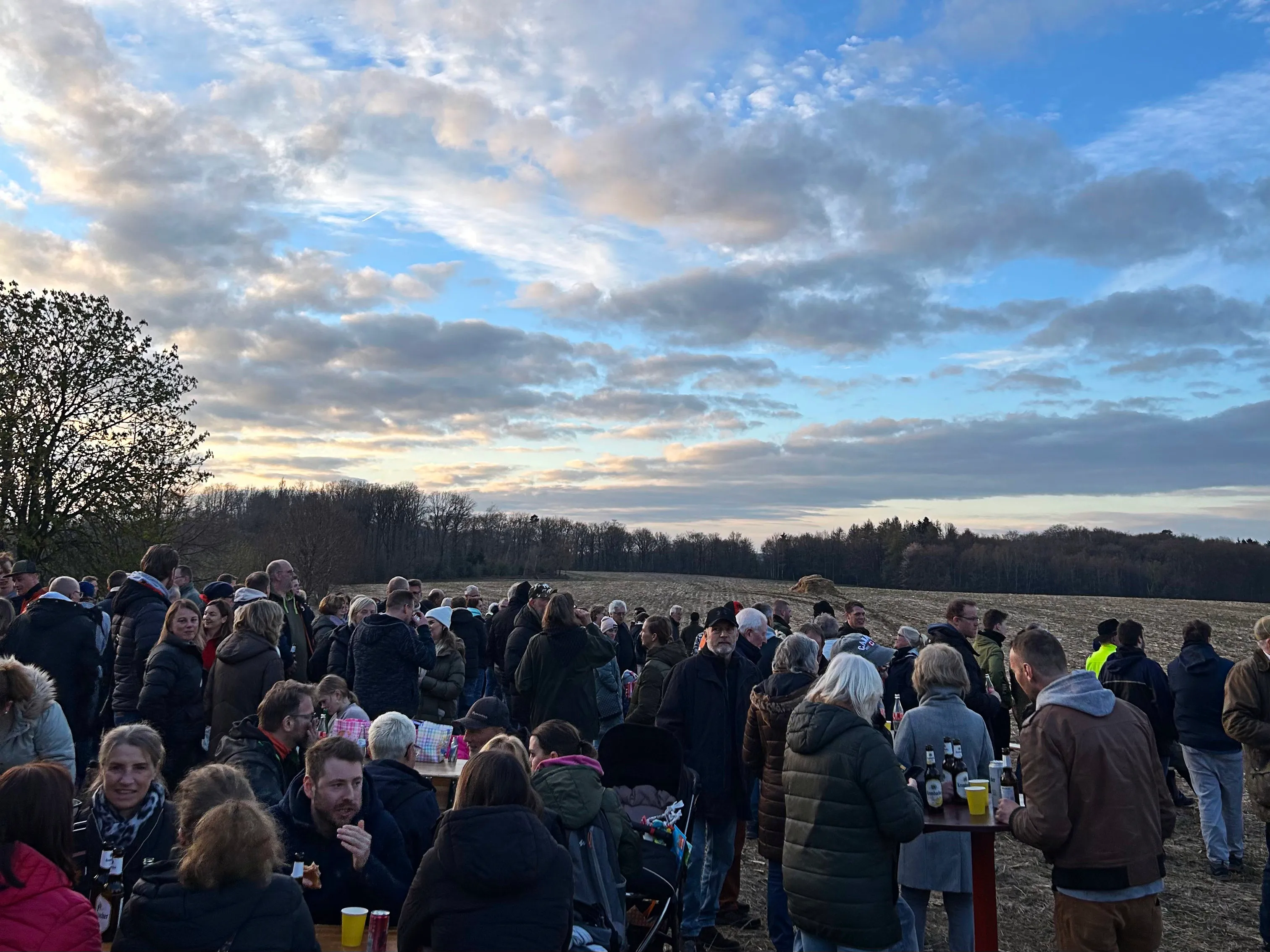 Große Menschenmenge vor einem brennenden Osterfeuer auf einem Feld, im Hintergrund Bäume und ein bewölkter Himmel.