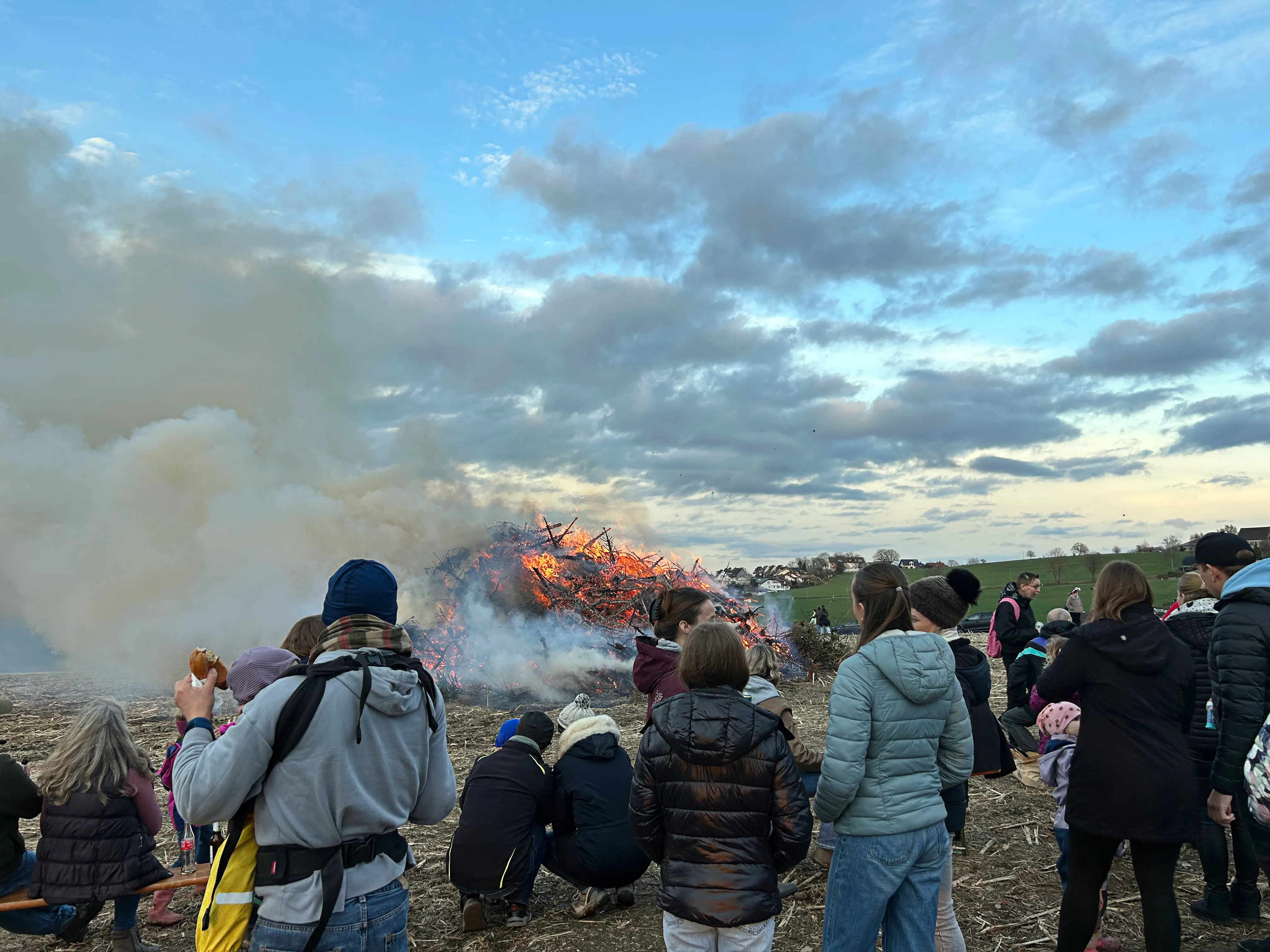 Menschen beobachten ein großes Osterfeuer mit Flammen und viel Rauch am Abend.