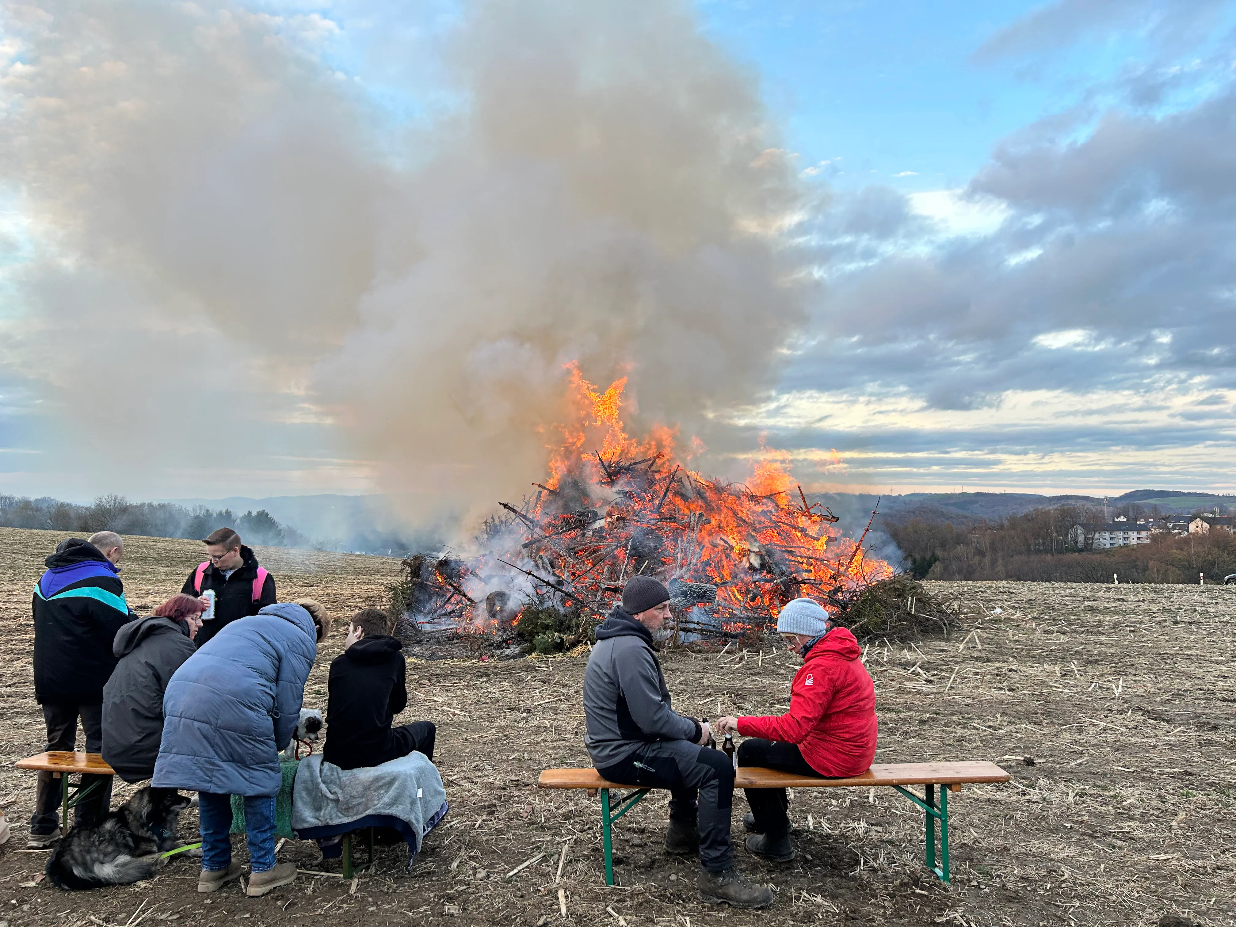 Großes Osterfeuer mit Flammen und Rauchwolken, umgeben von Menschen auf Bänken und im Stehen auf einem Feld.