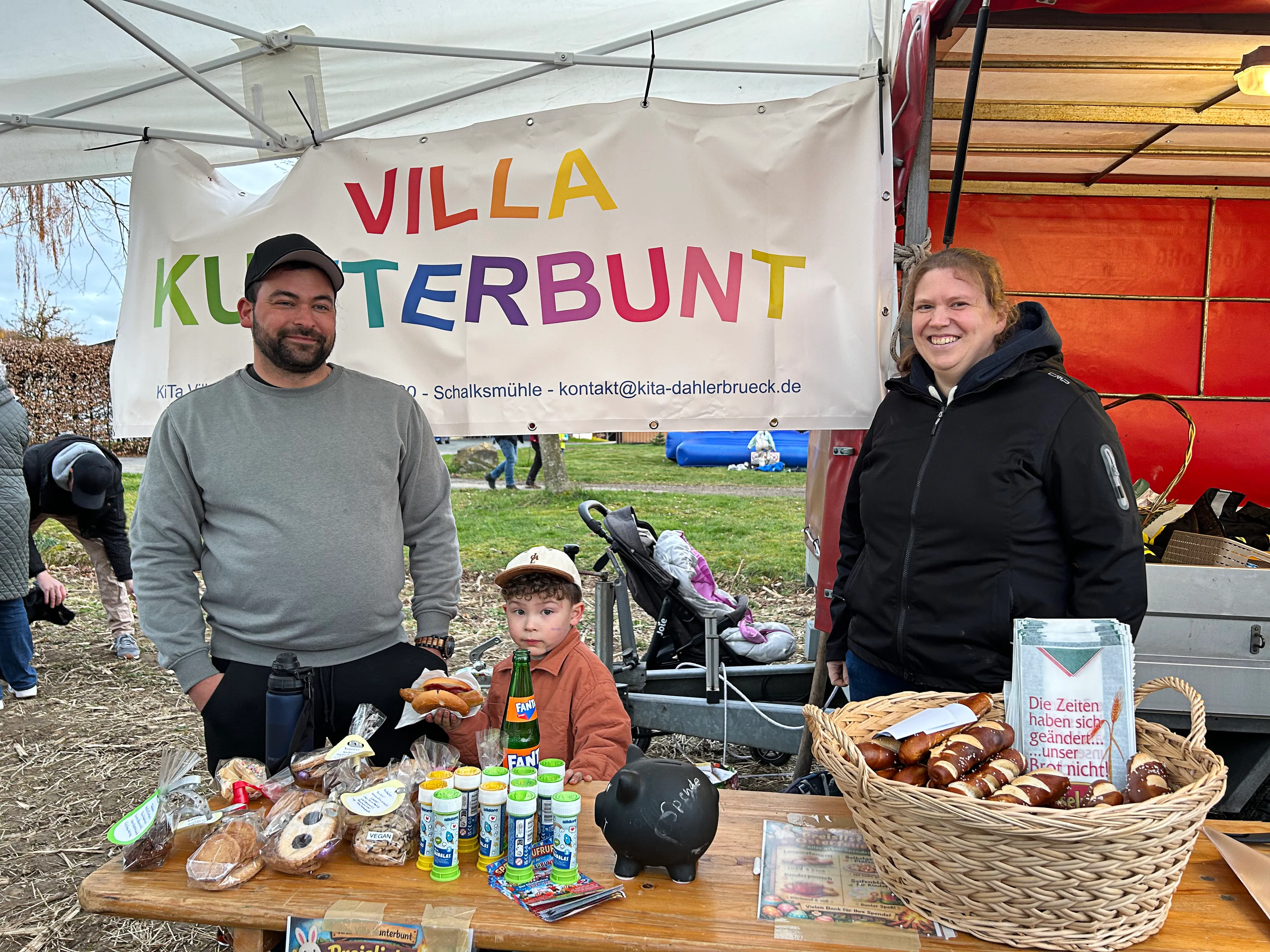 Ein Mann mit Bart, ein Kind und eine Frau stehen an einem Tisch mit Lebensmitteln vor einem bunten Banner mit der Aufschrift „Villa Kunterbunt“.