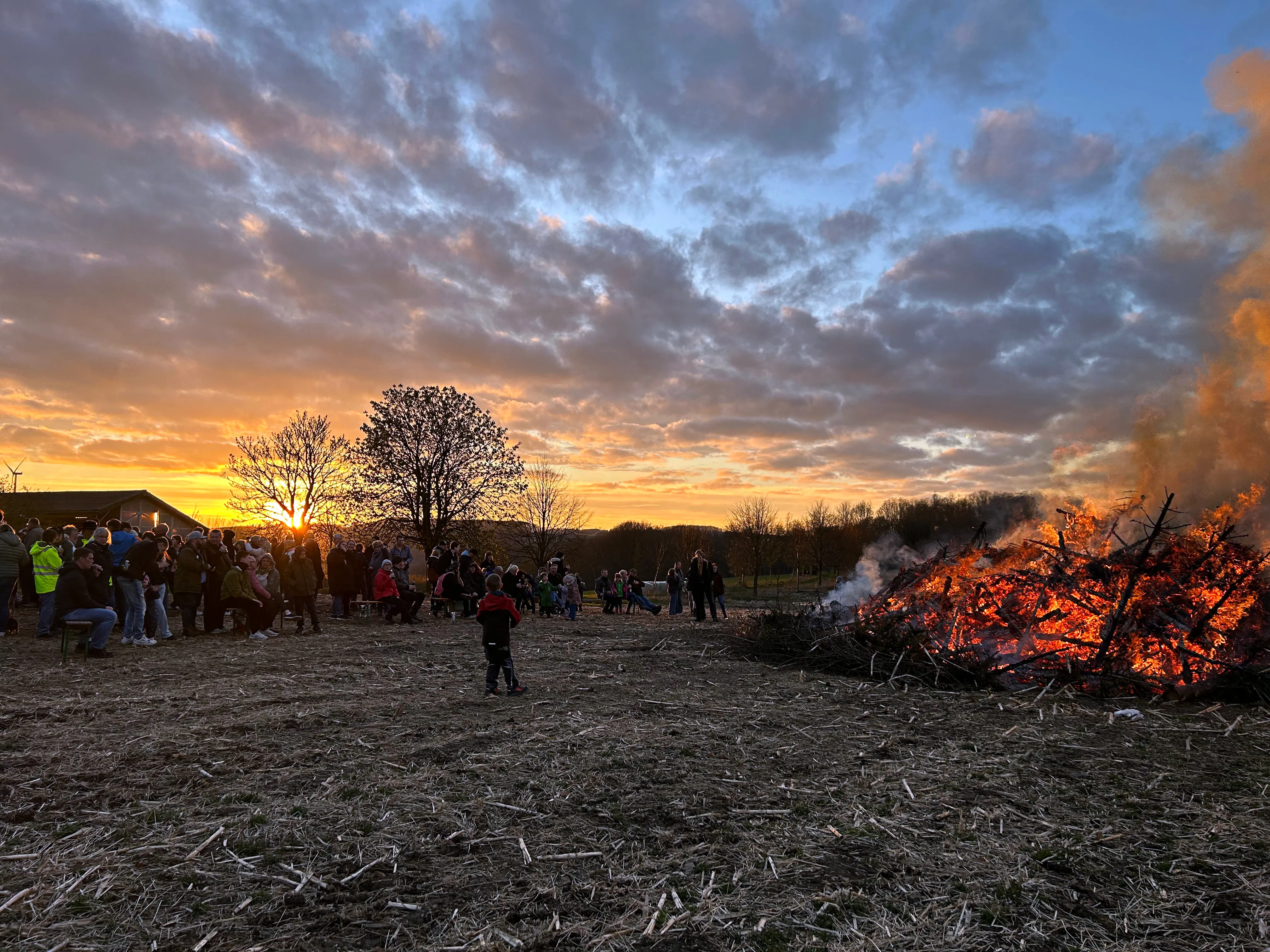 Großes Osterfeuer brennt auf einem Feld, umgeben von zahlreichen Zuschauern bei Sonnenuntergang.