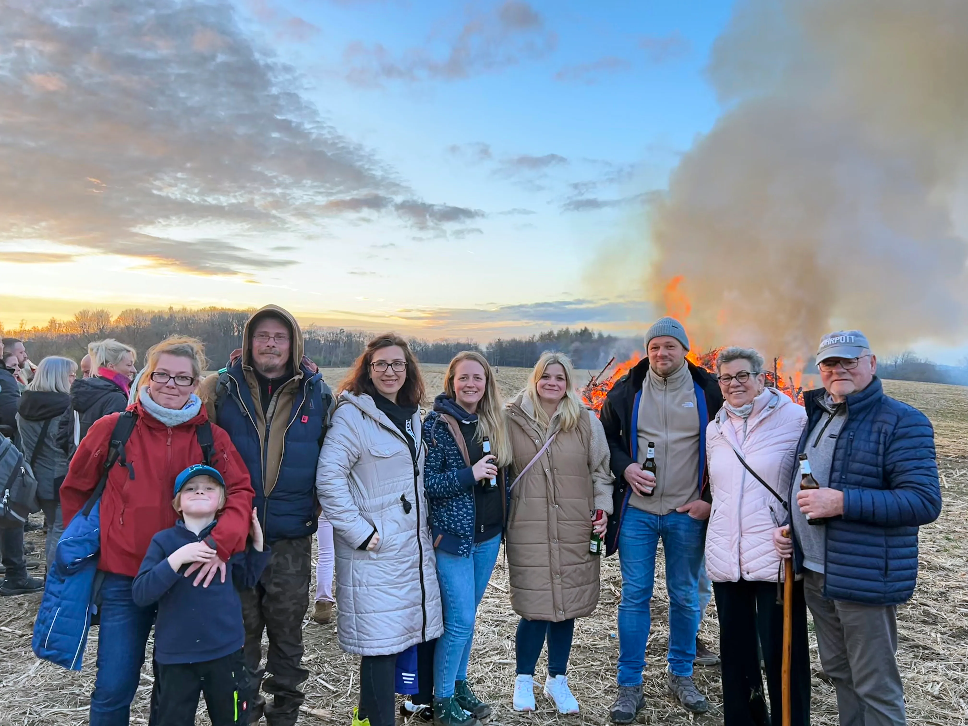 Eine Gruppe von Menschen steht vor einem großen Osterfeuer auf einem Feld, Rauch steigt auf. Einige tragen Mützen und Jacken, ein Kind hat eine Maske auf.