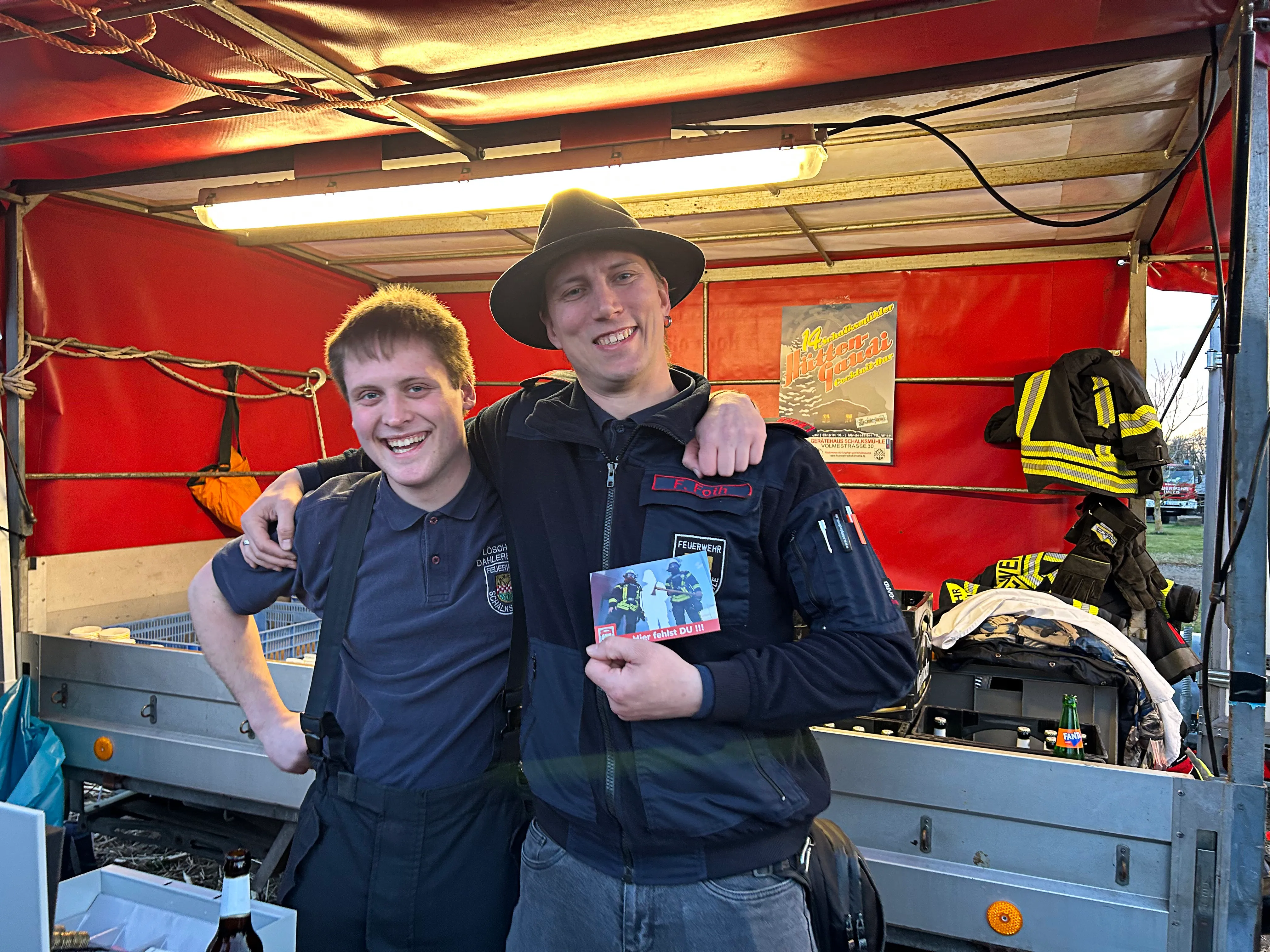 Zwei lächelnde Feuerwehrleute stehen eng nebeneinander und halten ein kleines Foto hoch. Im Hintergrund ist ein roter Verkaufsstand und weitere Einsatzkräfte zu sehen.