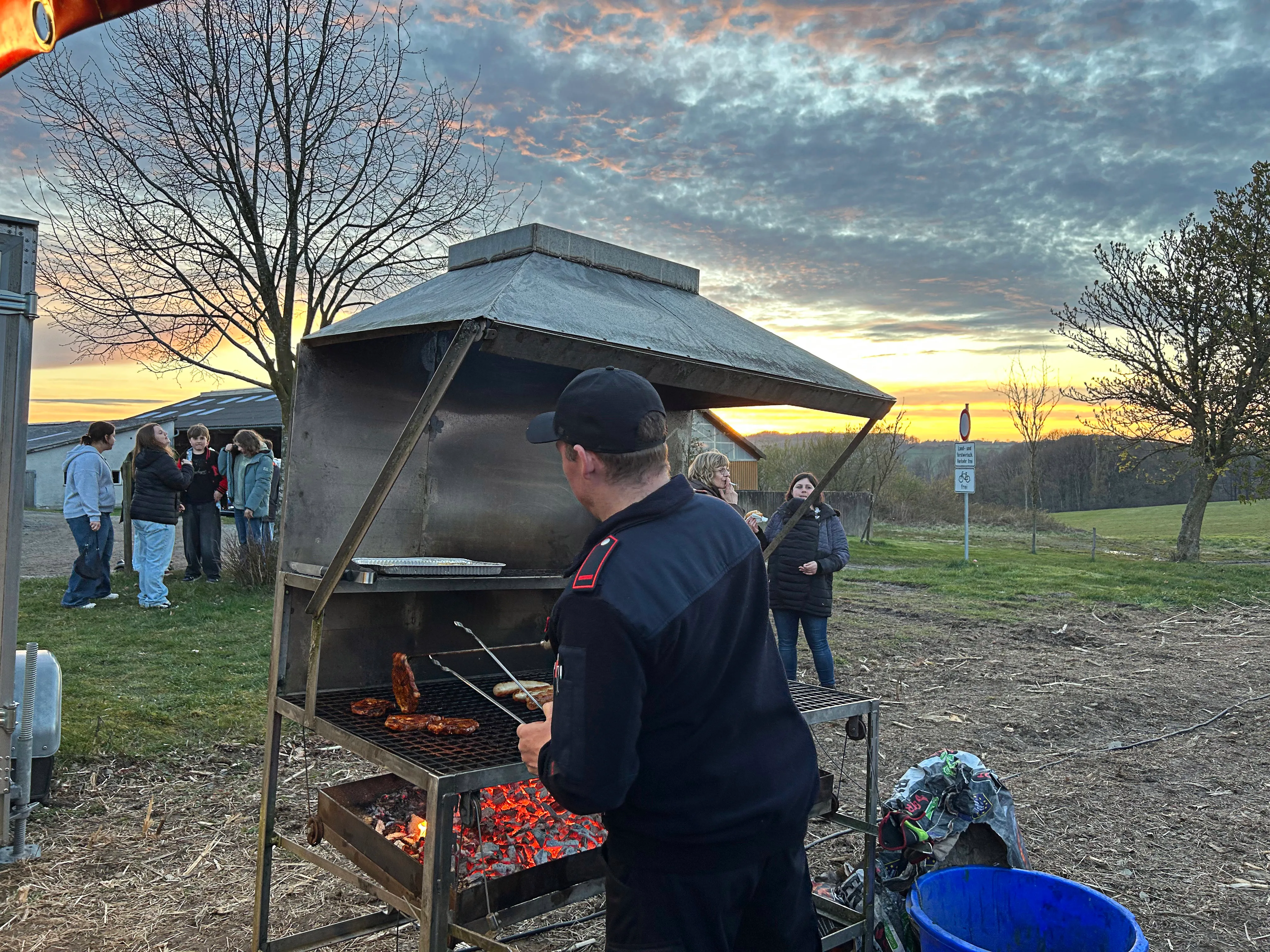 Ein Mann steht an einem großen Grill und bereitet Speisen zu, während im Hintergrund Menschen stehen und ein Baumwipfel zu sehen ist. Im Hintergrund ein Abendhimmel.