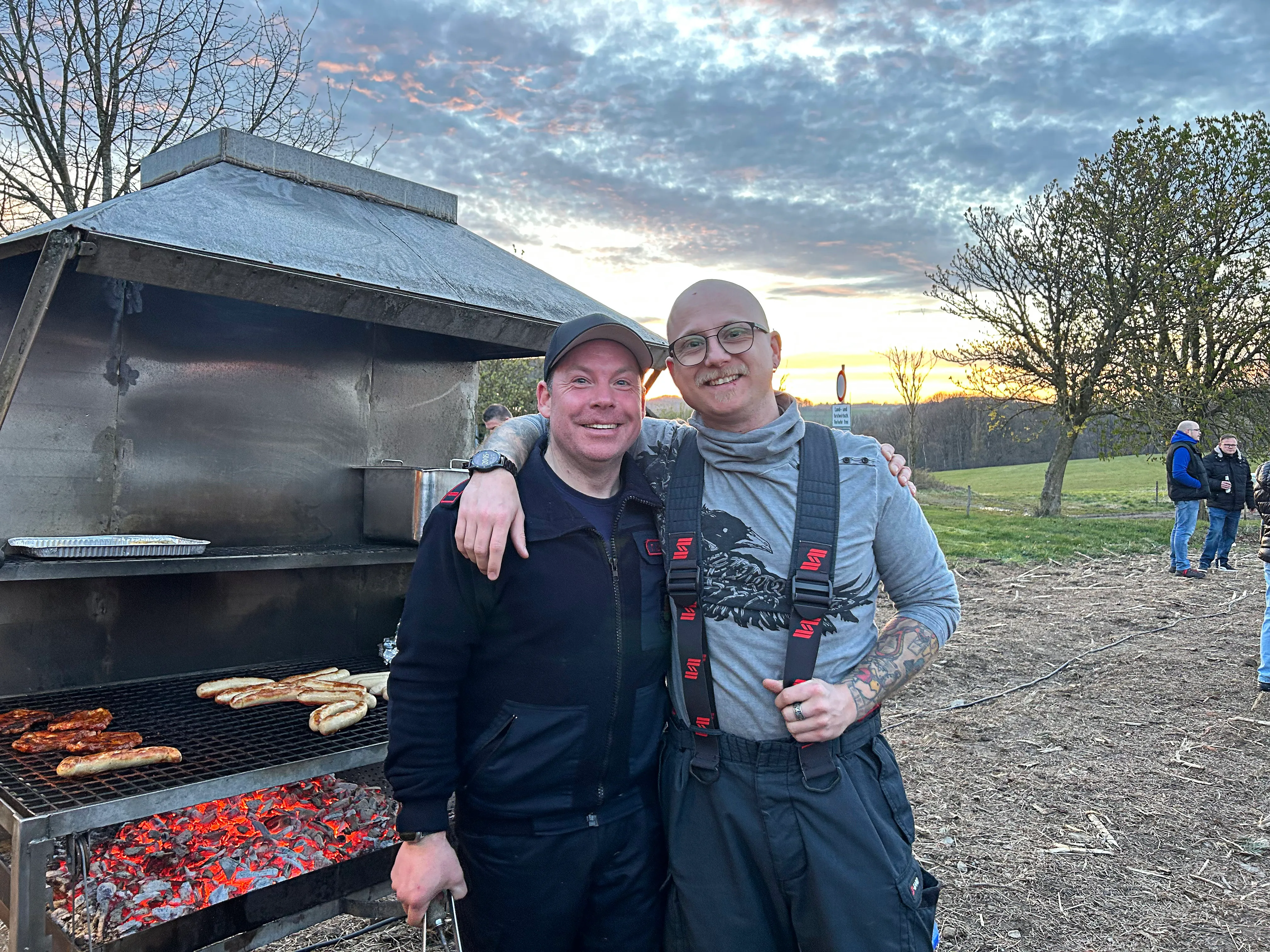 Zwei lächelnde Männer stehen vor einem großen Grill mit glühenden Kohlen und darauf liegendem Grillgut. Im Hintergrund ein Osterfeuer und weitere Personen.