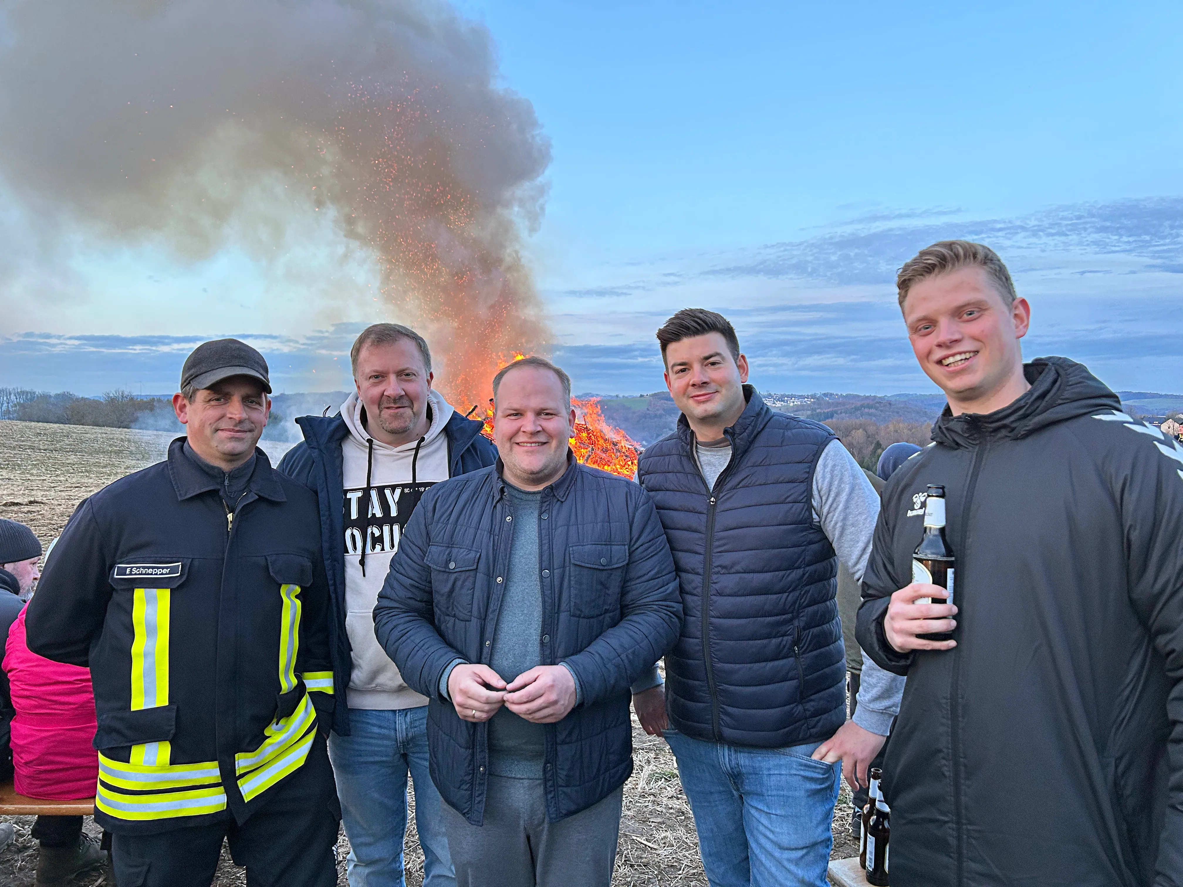 Fünf Männer stehen vor einem großen Osterfeuer mit Rauchwolken am Himmel. Ein Mann in Feuerwehruniform ist links im Bild zu sehen.