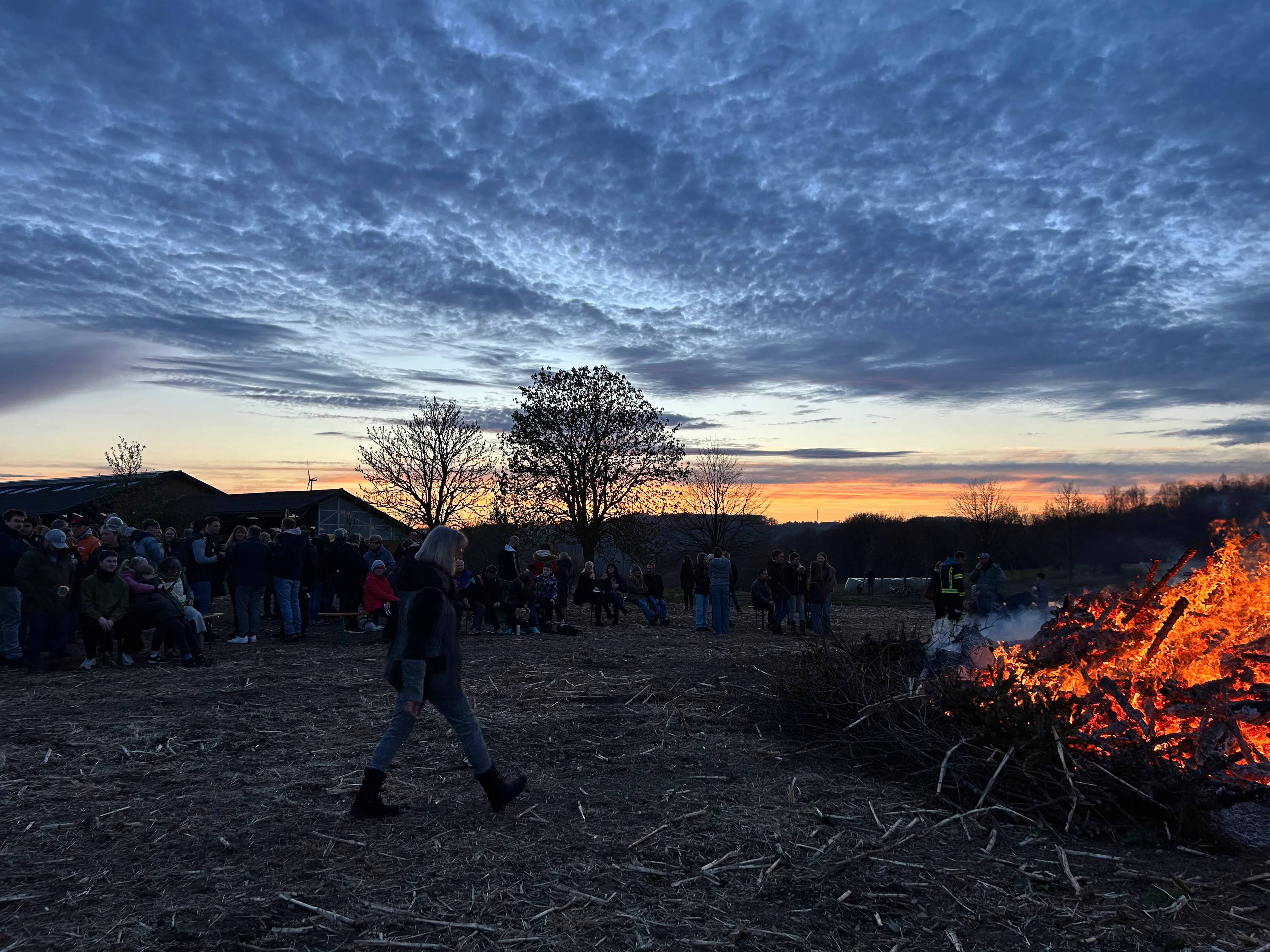 Großes Osterfeuer mit vielen Zuschauern im Abendlicht. Eine Person geht im Vordergrund auf das Feuer zu.