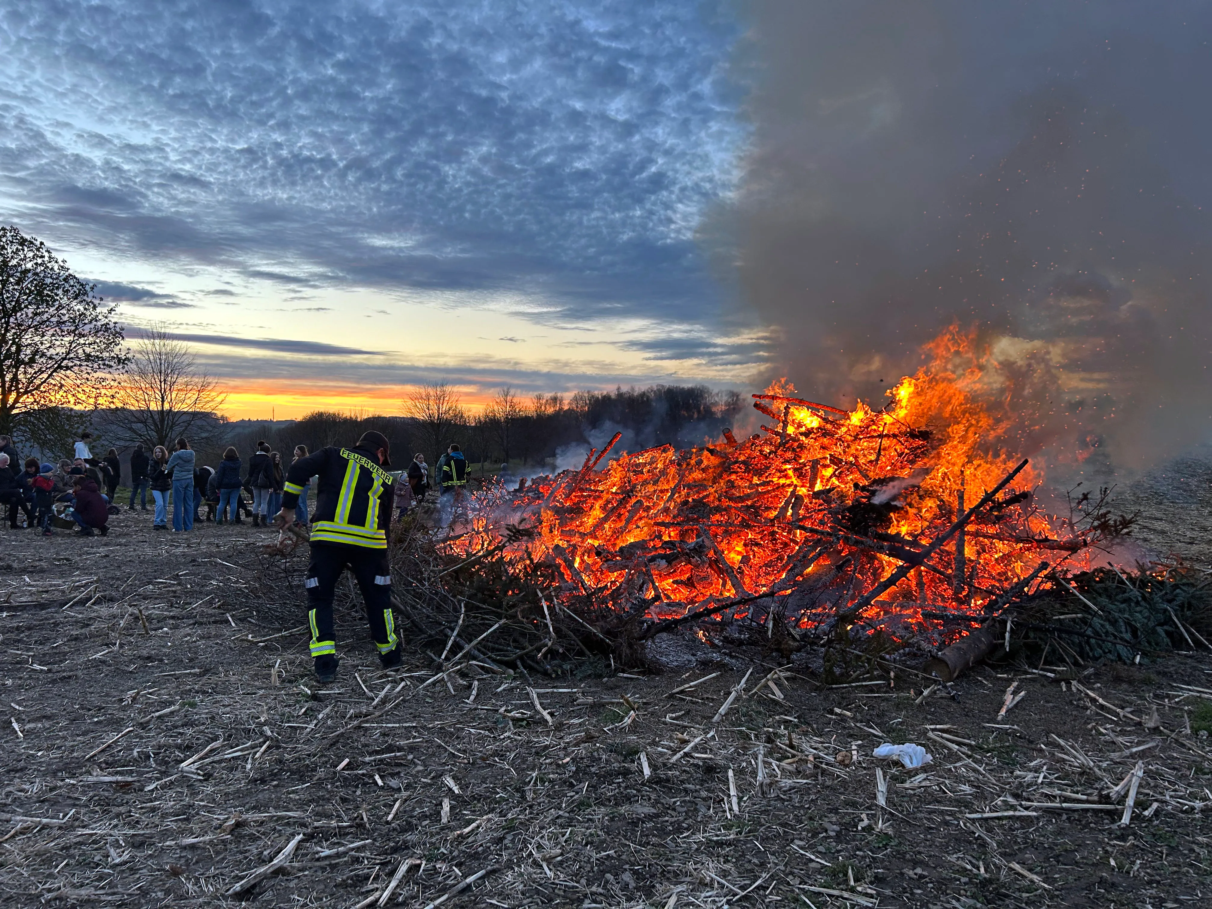 Großes Osterfeuer mit Flammen und Rauch vor einer Menschenmenge bei Sonnenuntergang.