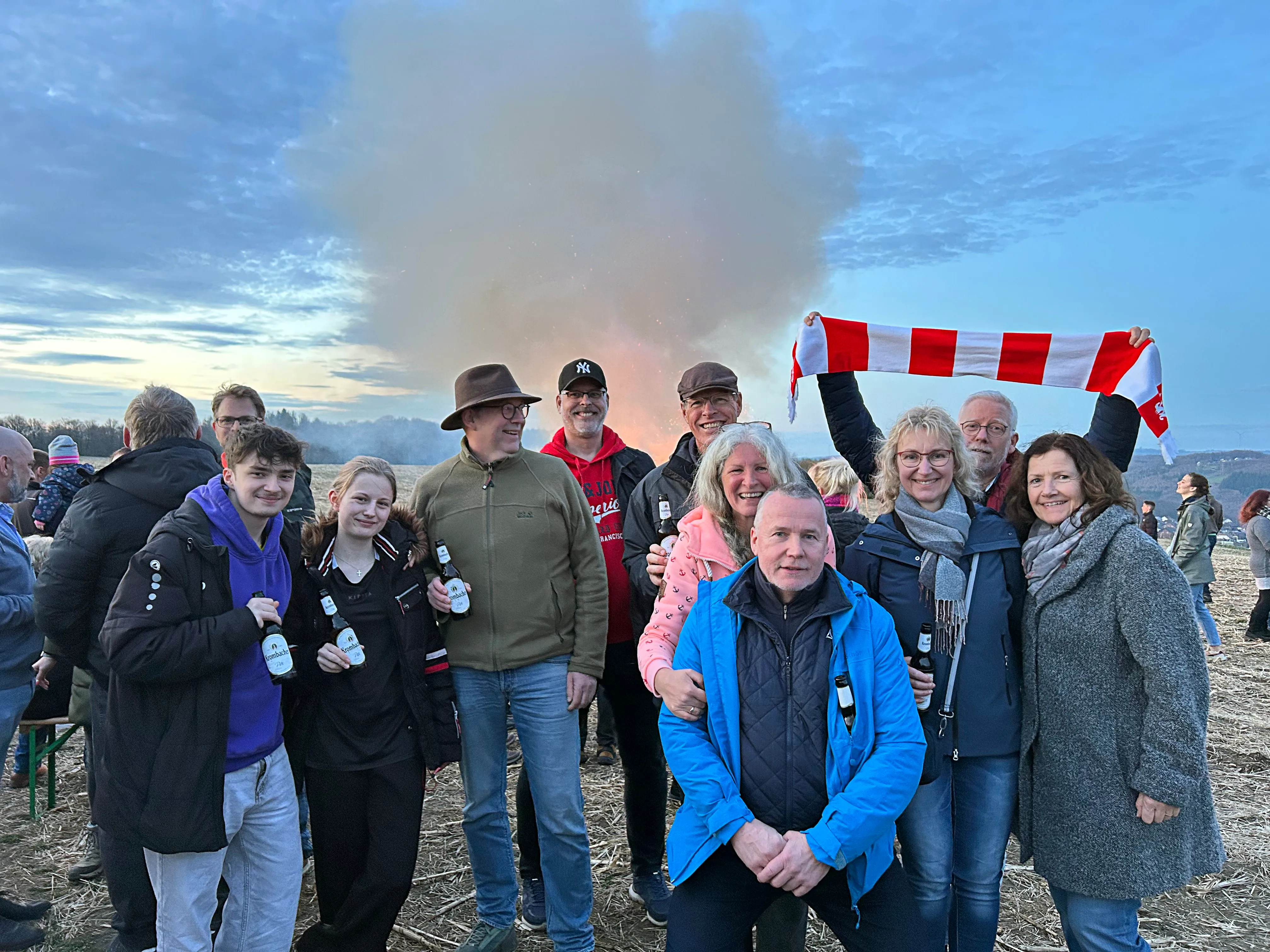 Eine Gruppe von Menschen steht vor einem großen Osterfeuer mit Rauchfahnen und hält Bierflaschen in den Händen. Im Hintergrund weht eine rot-weiß gestreifte Fahne.
