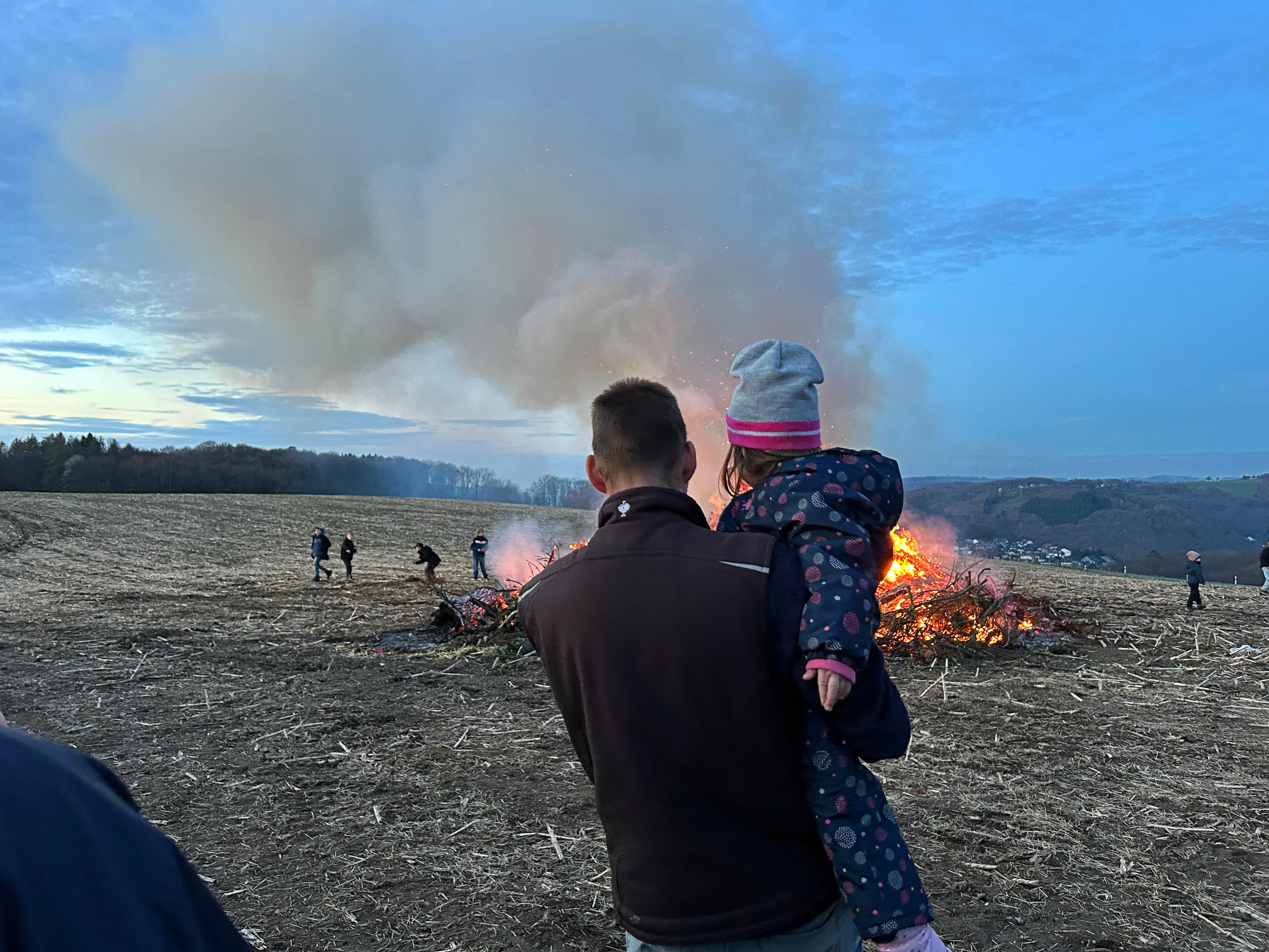 Ein Mann trägt ein Kind auf seinen Schultern, während im Hintergrund ein großes Osterfeuer brennt und Menschen stehen.