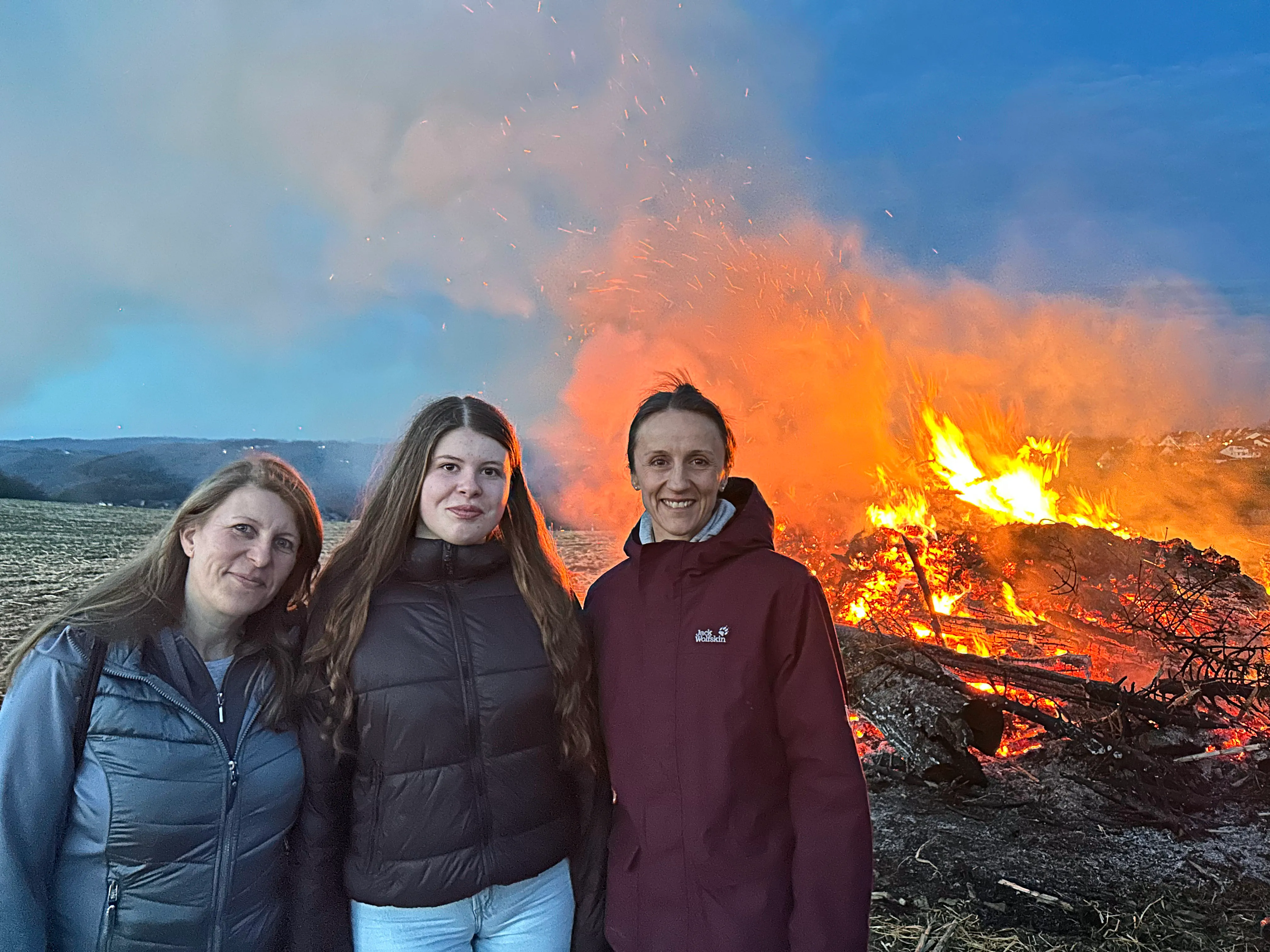 Drei junge Frauen stehen vor einem großen Osterfeuer mit hellen Flammen und viel Rauch am Abend.