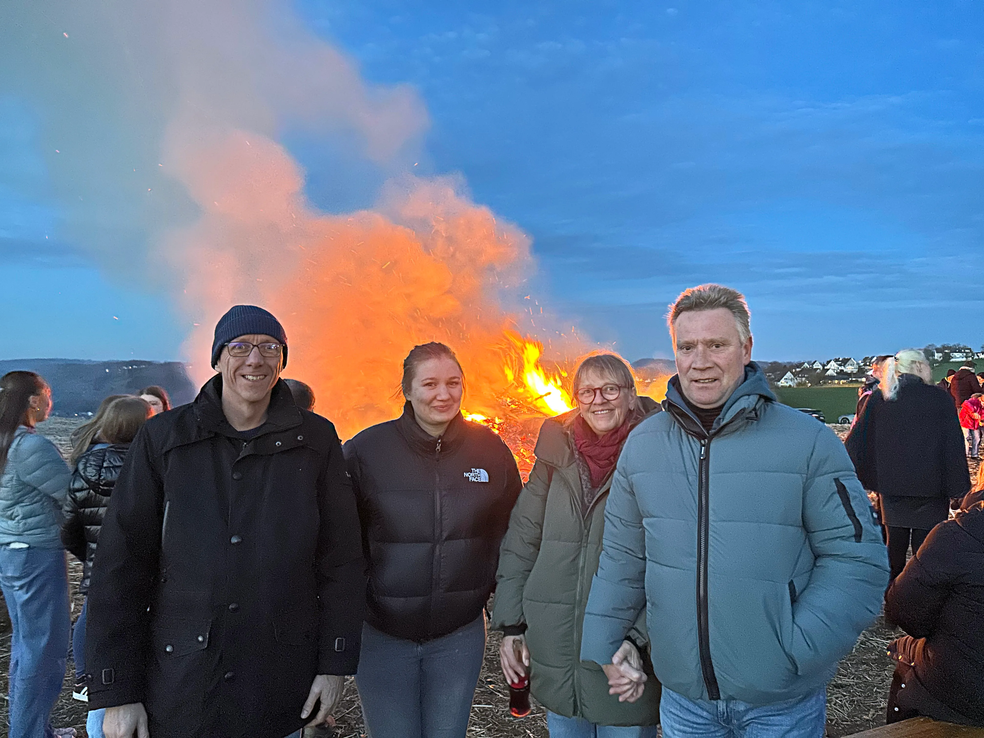 Fünf Personen stehen vor einem großen Osterfeuer, das hell leuchtet und Rauchwolken aufwirft. Im Hintergrund sind weitere Menschen und Häuser zu sehen.