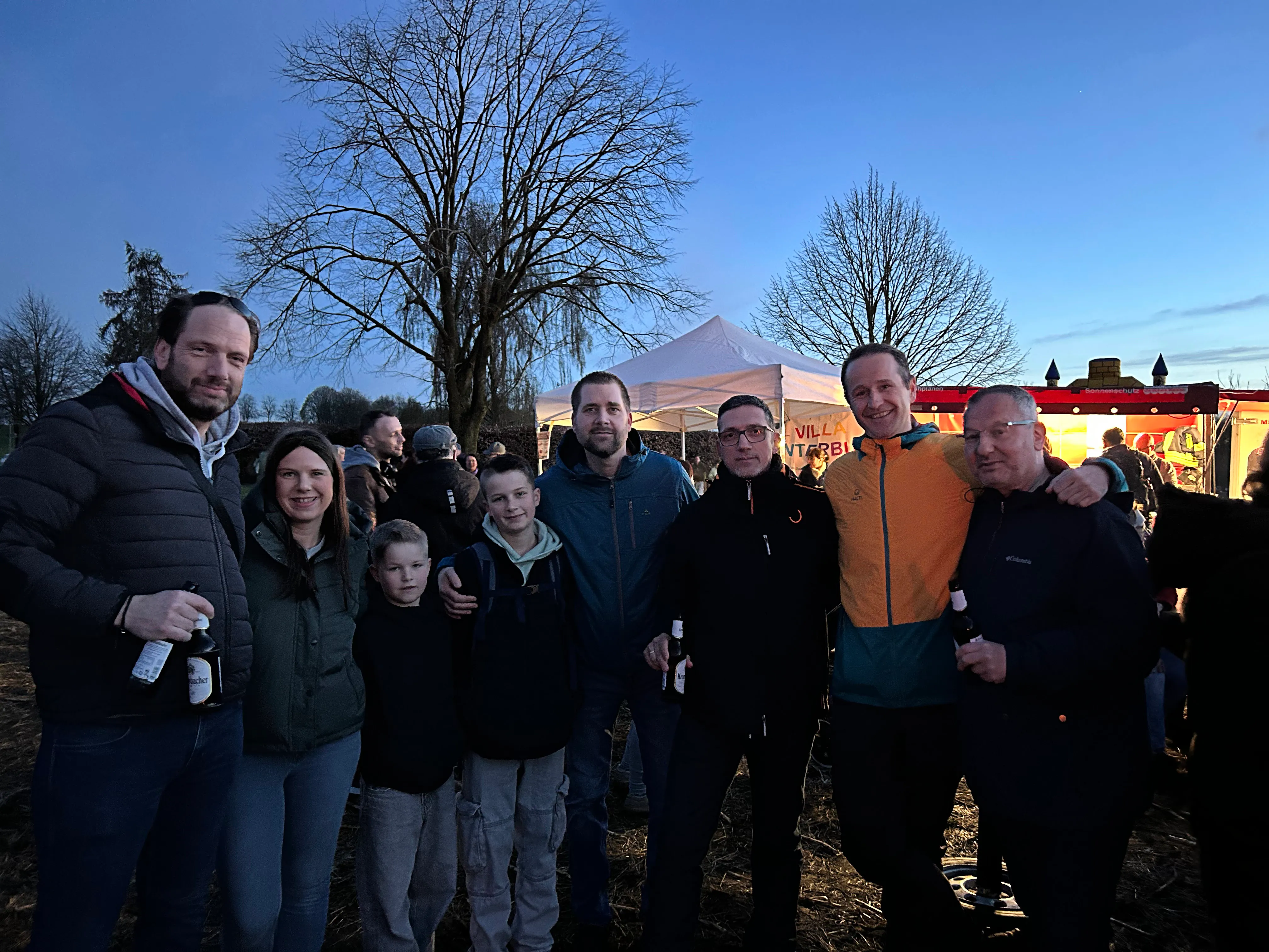 Eine Gruppe von Menschen steht zusammen vor einem großen Baum und einem weißen Zelt, im Hintergrund ein beleuchteter Wagen. Das Osterfeuer in Dahlerbrück lockte viele Besucher an.